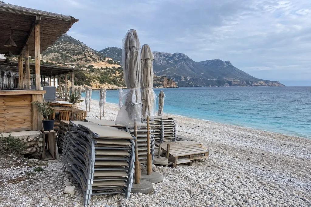 Stacked sun loungers and closed parasols on a pebble beach before the season, turquoise sea and mountains behind
